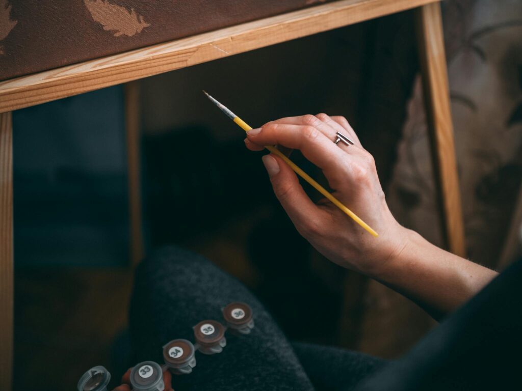 Close-up of an artist's hand holding a paintbrush while painting on canvas indoors.